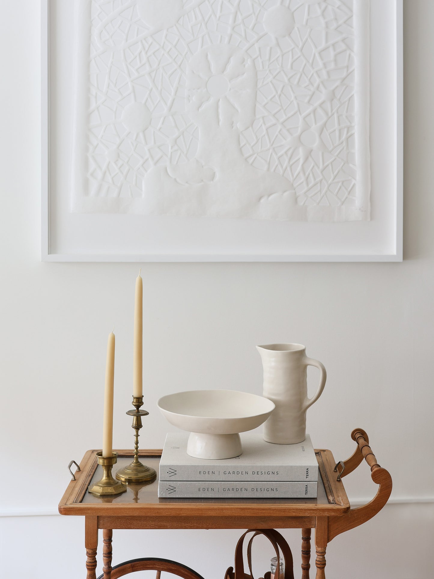 BAR CART on a wooden wheeled stand with books, a ceramic bowl, pitcher, and brass candlesticks beside white wall art.