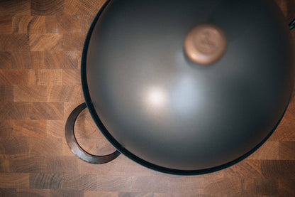 Close-up view of a domed carbon steel bread cloche lid resting on a wooden butcher block countertop, featuring a copper handle accent and a metal handle grip.