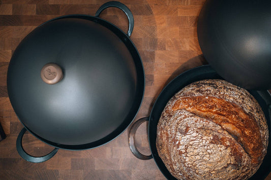 Top-down view of a dark bread cloche lid on carbon steel baking pan and rustic country loaf with cracked crust.