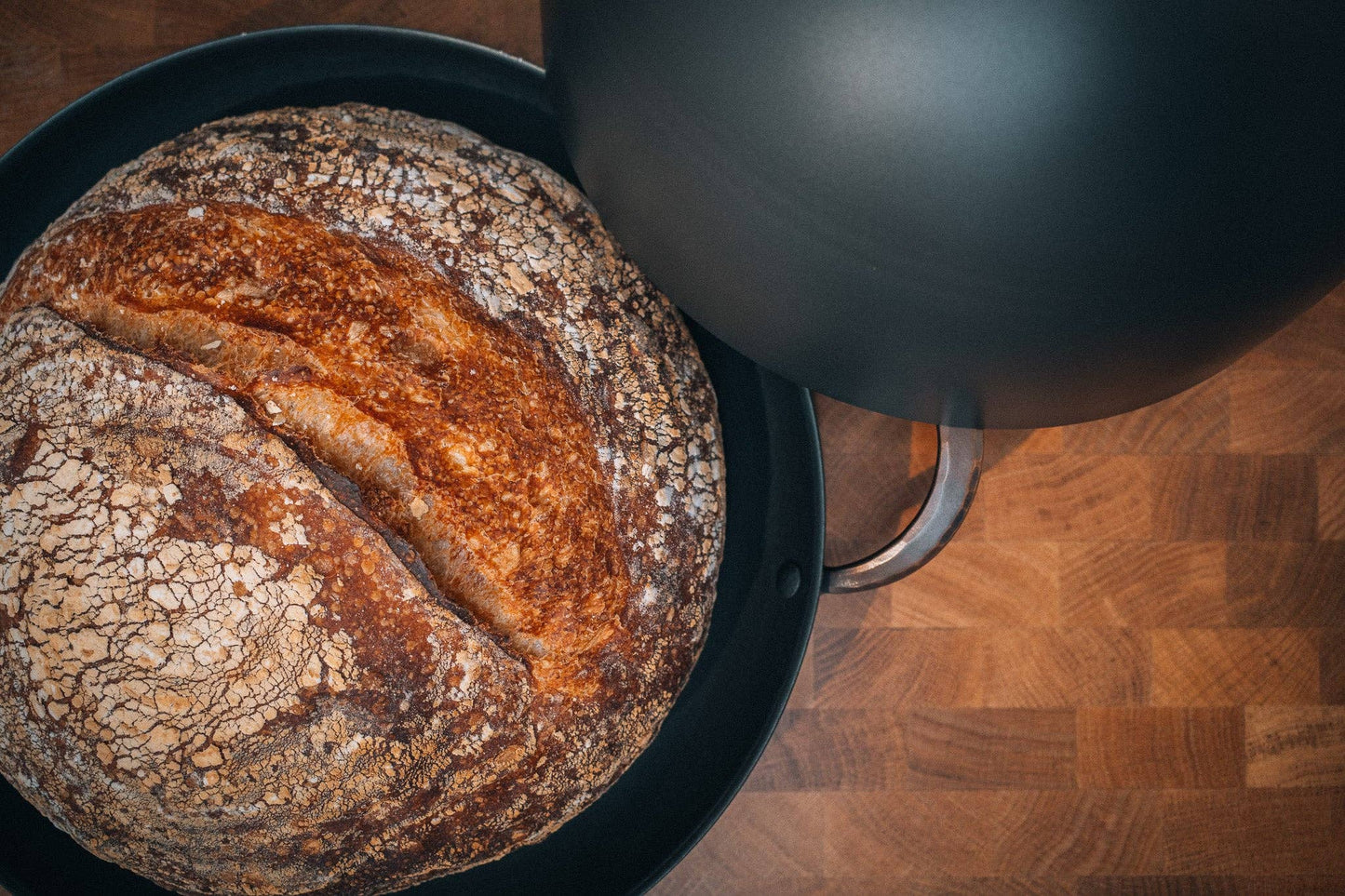 Rustic bread boule baked in 11-inch roaster revealed by lifted carbon steel cloche lid.